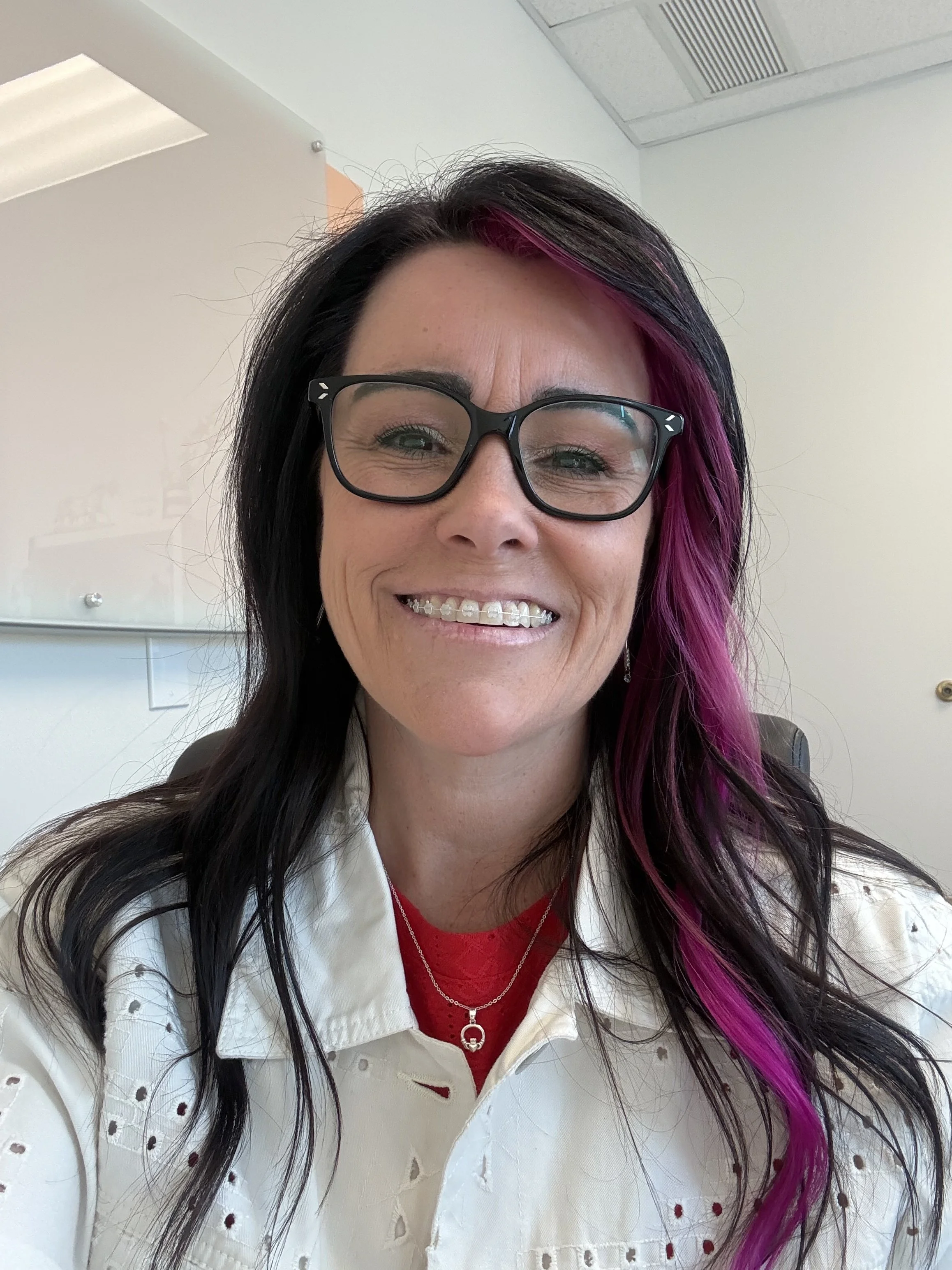 A woman with long black hair with purple streaks, wearing glasses, a white eyelet jacket, a red shirt, and a silver necklace with a heart-shaped pendant, smiling.