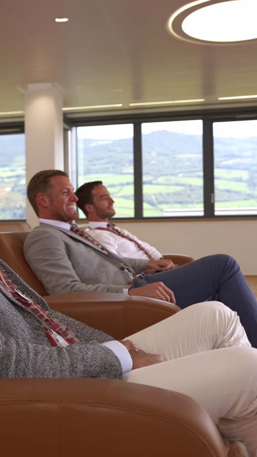 Three men sitting on leather chairs in a modern office or lounge area, looking out the large windows at a scenic landscape of mountains and green fields.