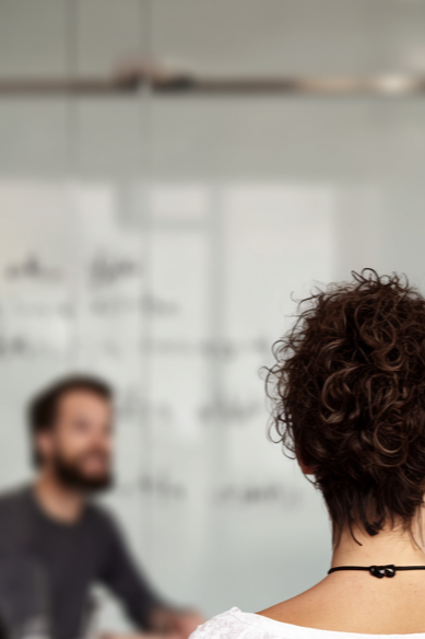 Back of a woman's head with curly hair in a classroom or office, with a man blurred in the background.