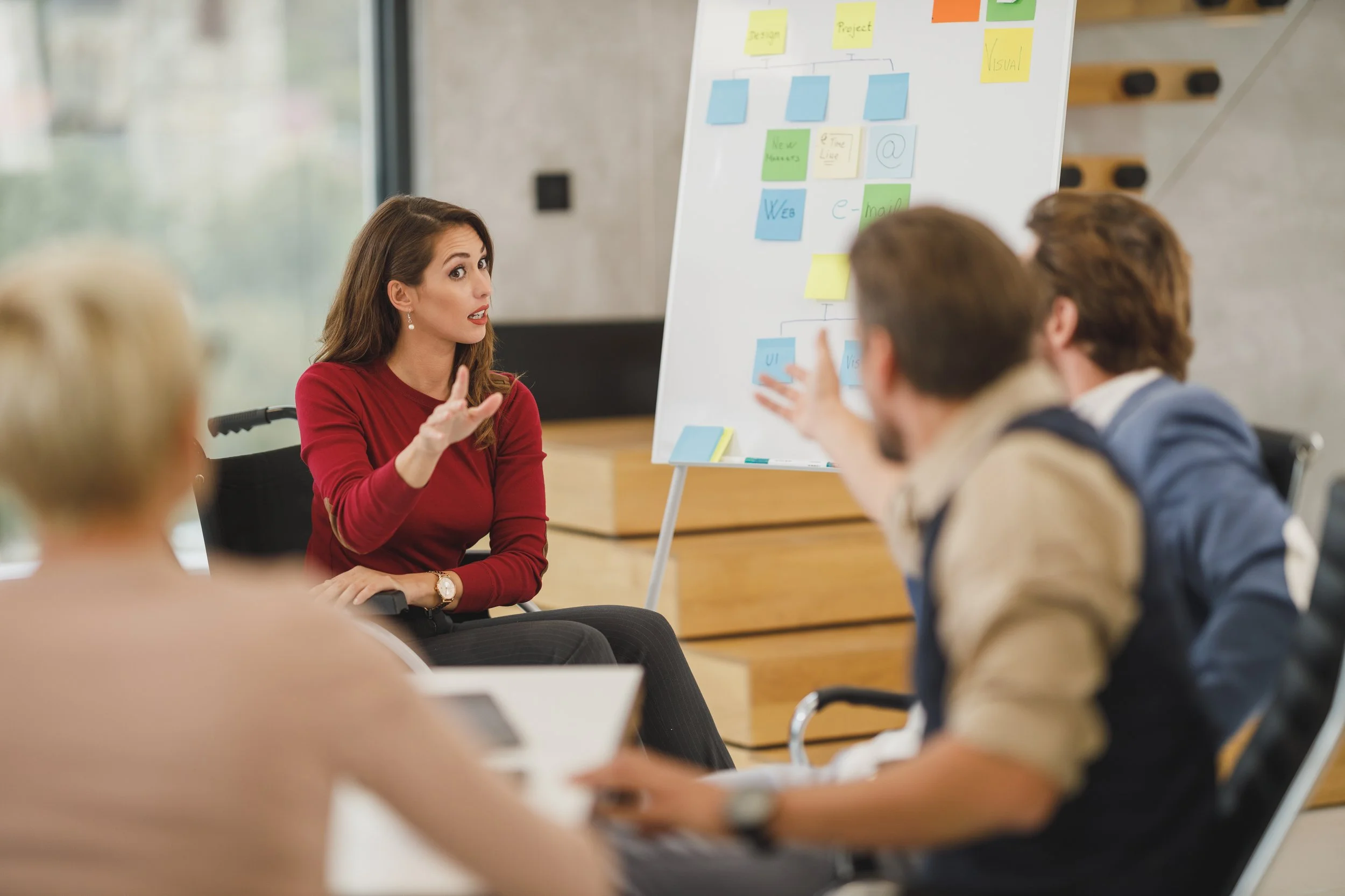 A woman in a red top is speaking to a group of people during a meeting, with a whiteboard filled with colorful sticky notes and diagrams in the background.
