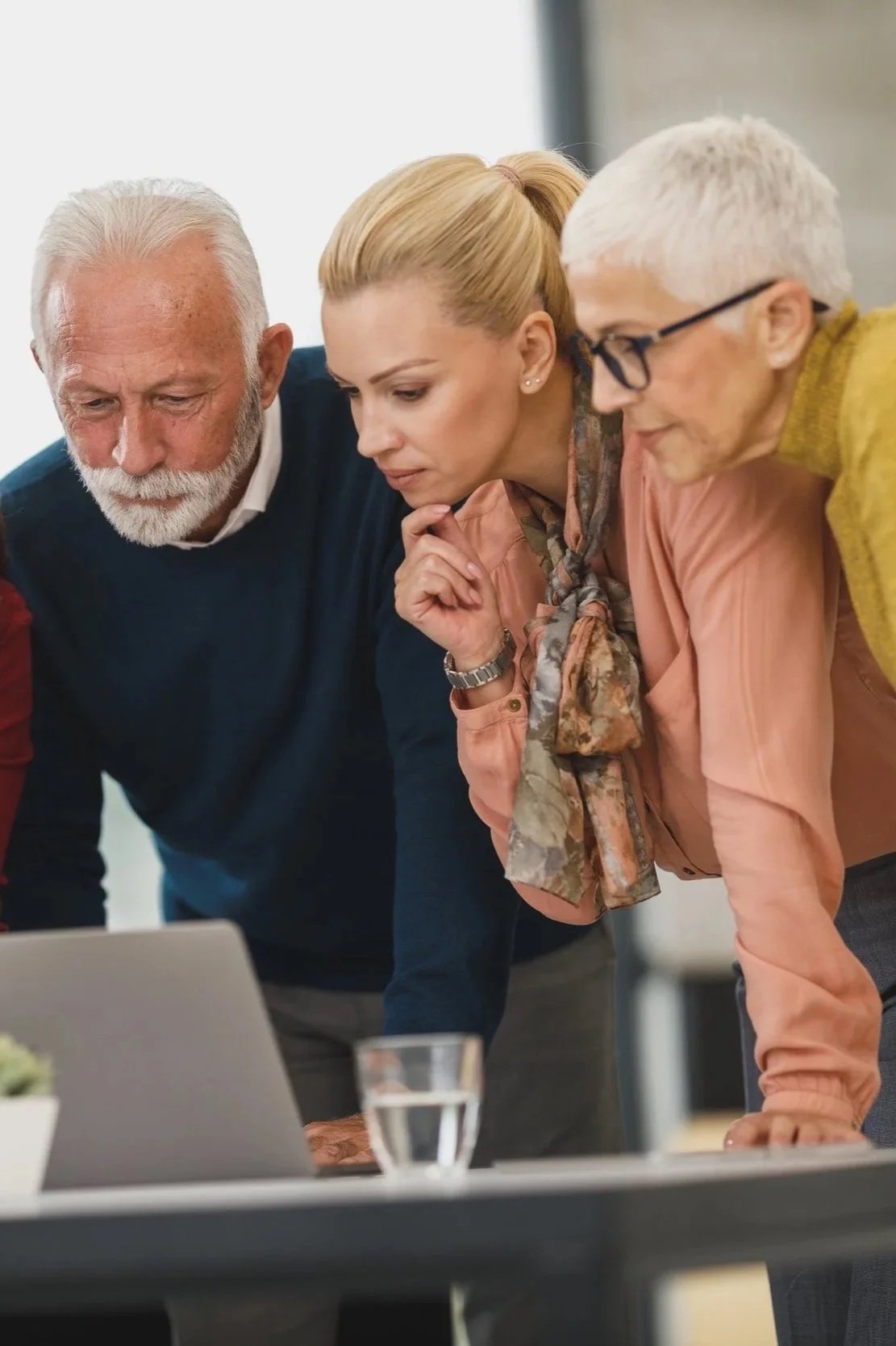 Three older adults, two women and one man, are closely looking at a laptop screen in a modern office setting.