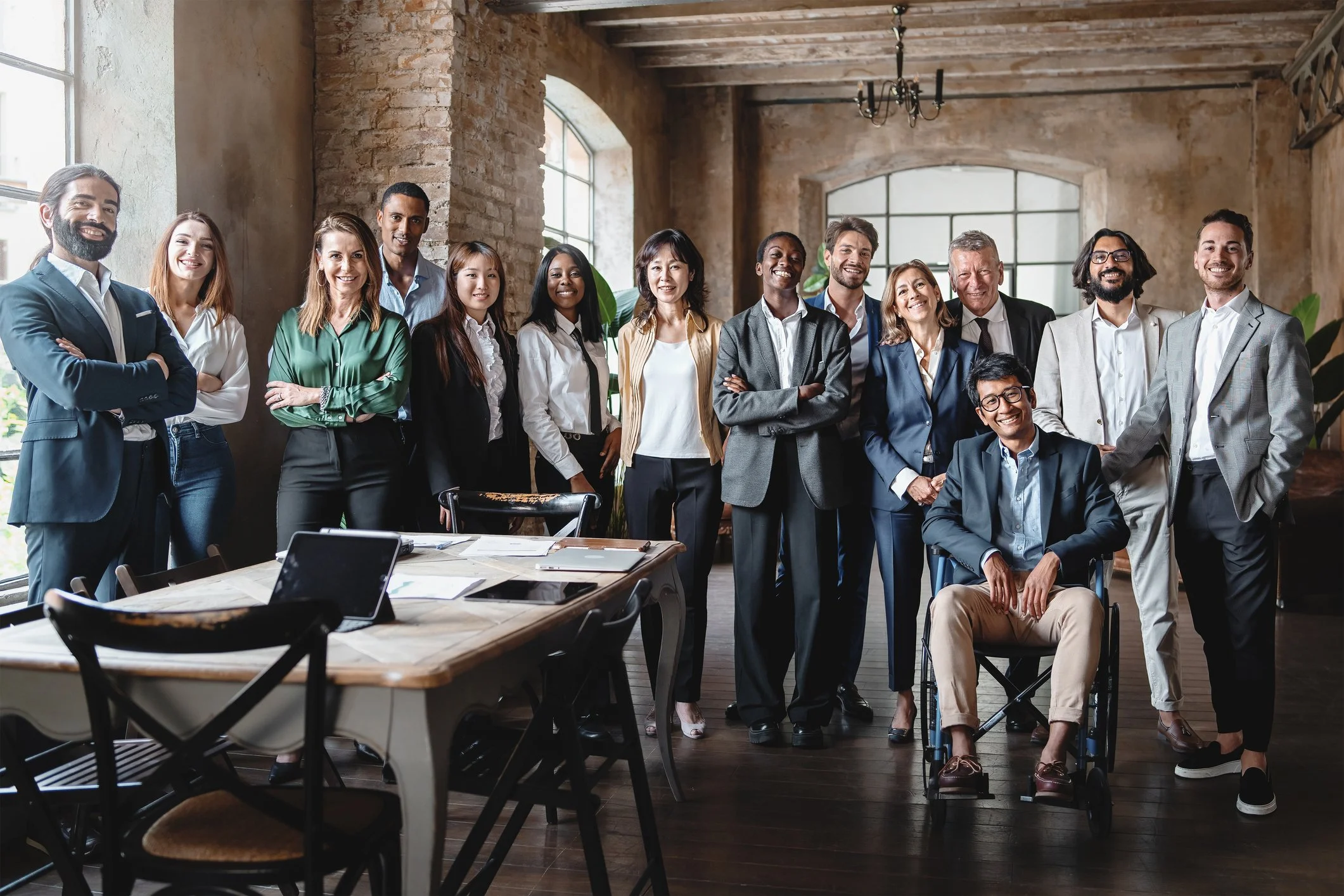 Group of diverse professionals standing and sitting in a spacious, rustic office with large windows and exposed brick walls.