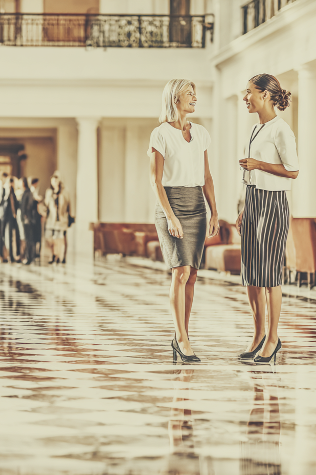 Two women are standing and talking in a bright, spacious lobby or atrium with a marble floor, surrounded by other people and seating areas.