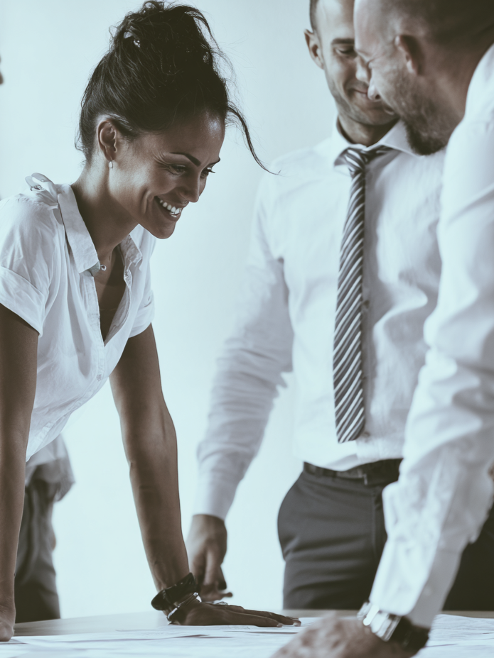 Three business professionals, two men and one woman, smiling and engaging in a discussion around a table in an office setting.