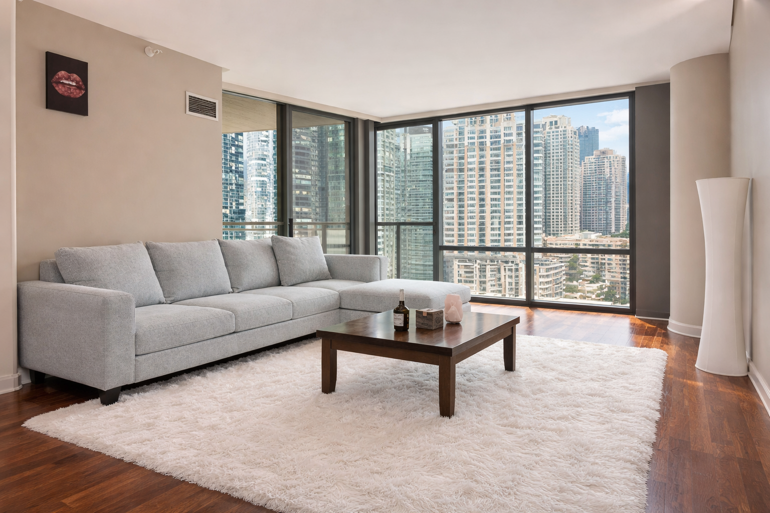 Modern living room with a light gray sofa, a wooden coffee table, and a large white shag rug, against floor-to-ceiling windows showing a city skyline.