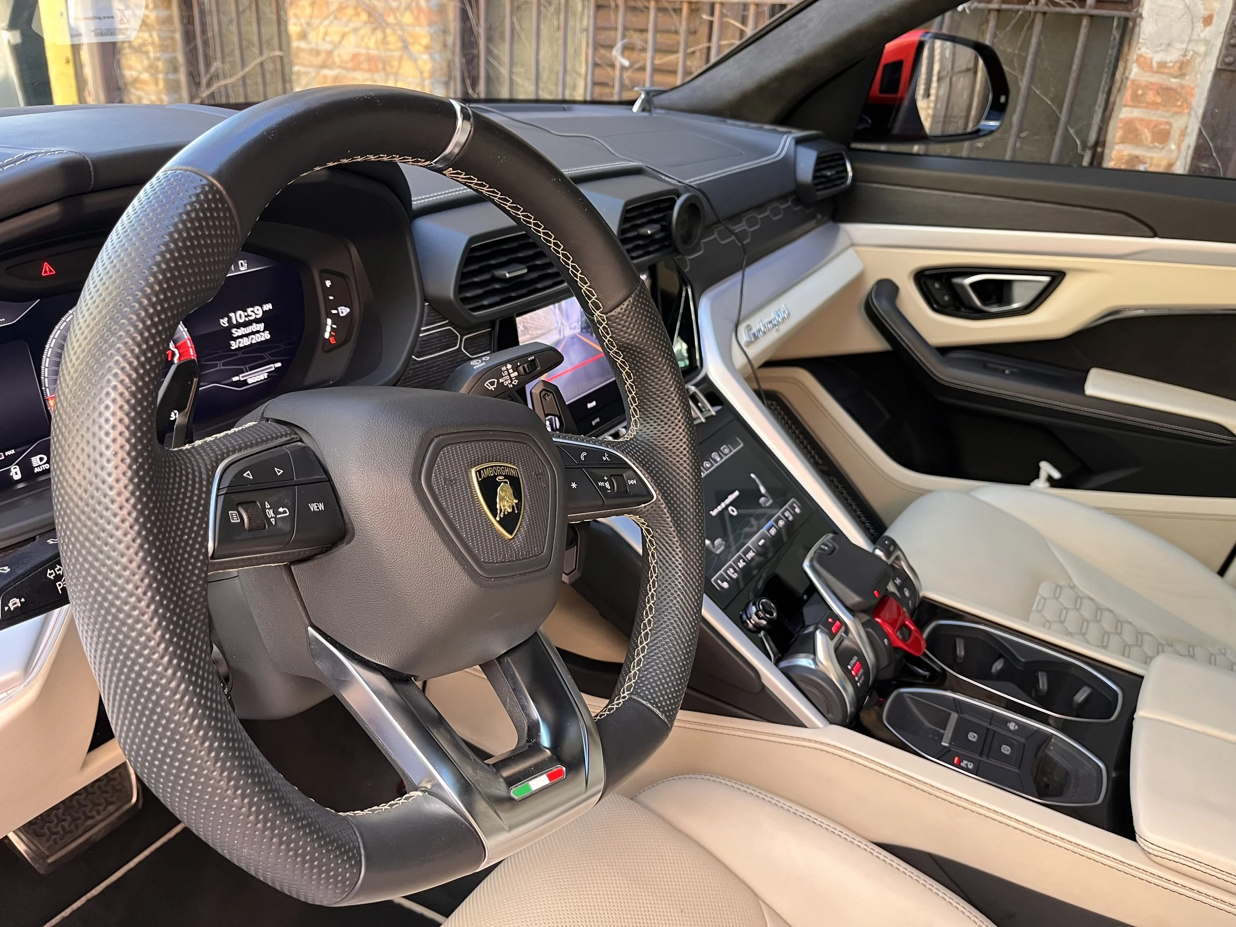 The interior of a modern luxury Lamborghini vehicle showing a steering wheel with the Lamborghini logo, digital dashboard, center console, and dashboard with air conditioning vents and digital displays, with a side mirror reflecting the outside envir