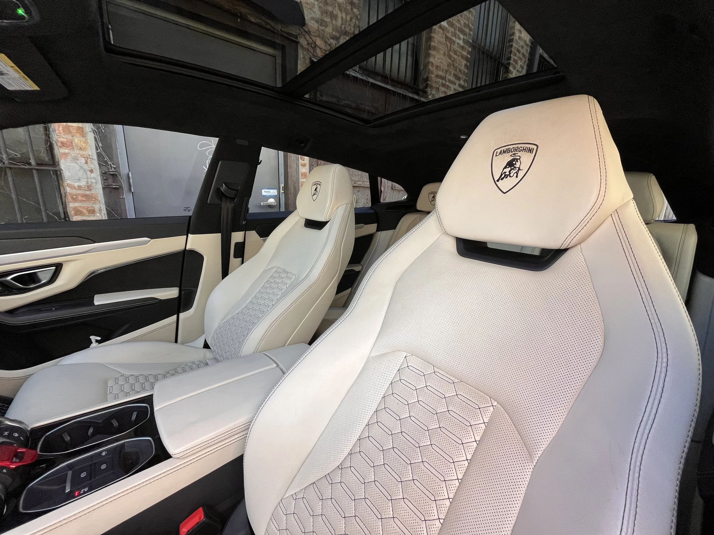 Interior of a luxury car with cream-colored leather seats embroidered with Lamborghini logos, and a panoramic sunroof showing neighboring brick buildings.