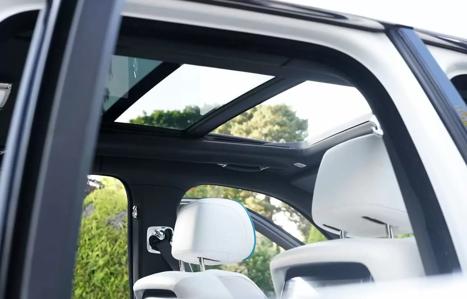 The interior of a modern vehicle with white leather seats and a panoramic sunroof, showing a clear sky and green trees outside.