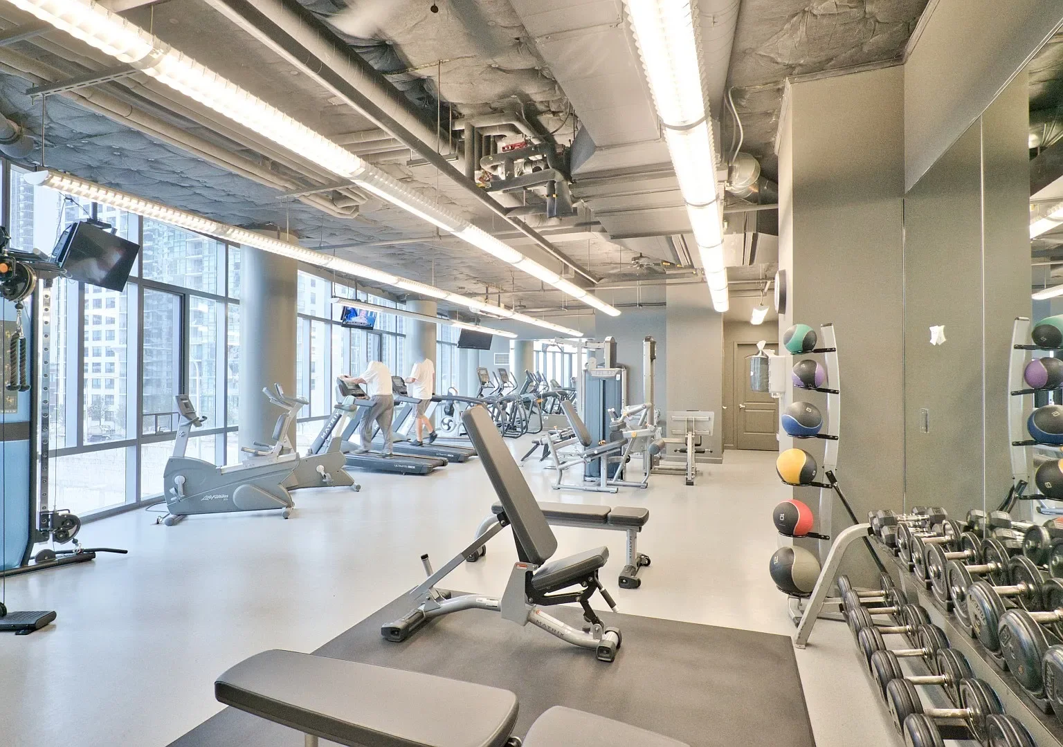 Interior of a modern gym with various cardio and weight lifting equipment, large windows showing city buildings outside, and people working out.