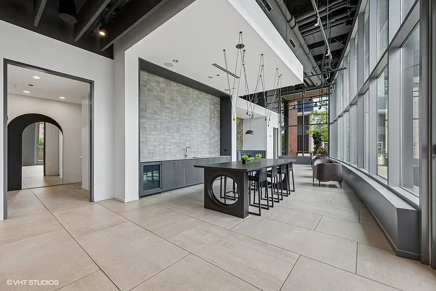Modern interior space with a large black black dining table, black chairs, and a kitchenette with a desk and sink, surrounded by floor-to-ceiling windows and contemporary lighting fixtures.