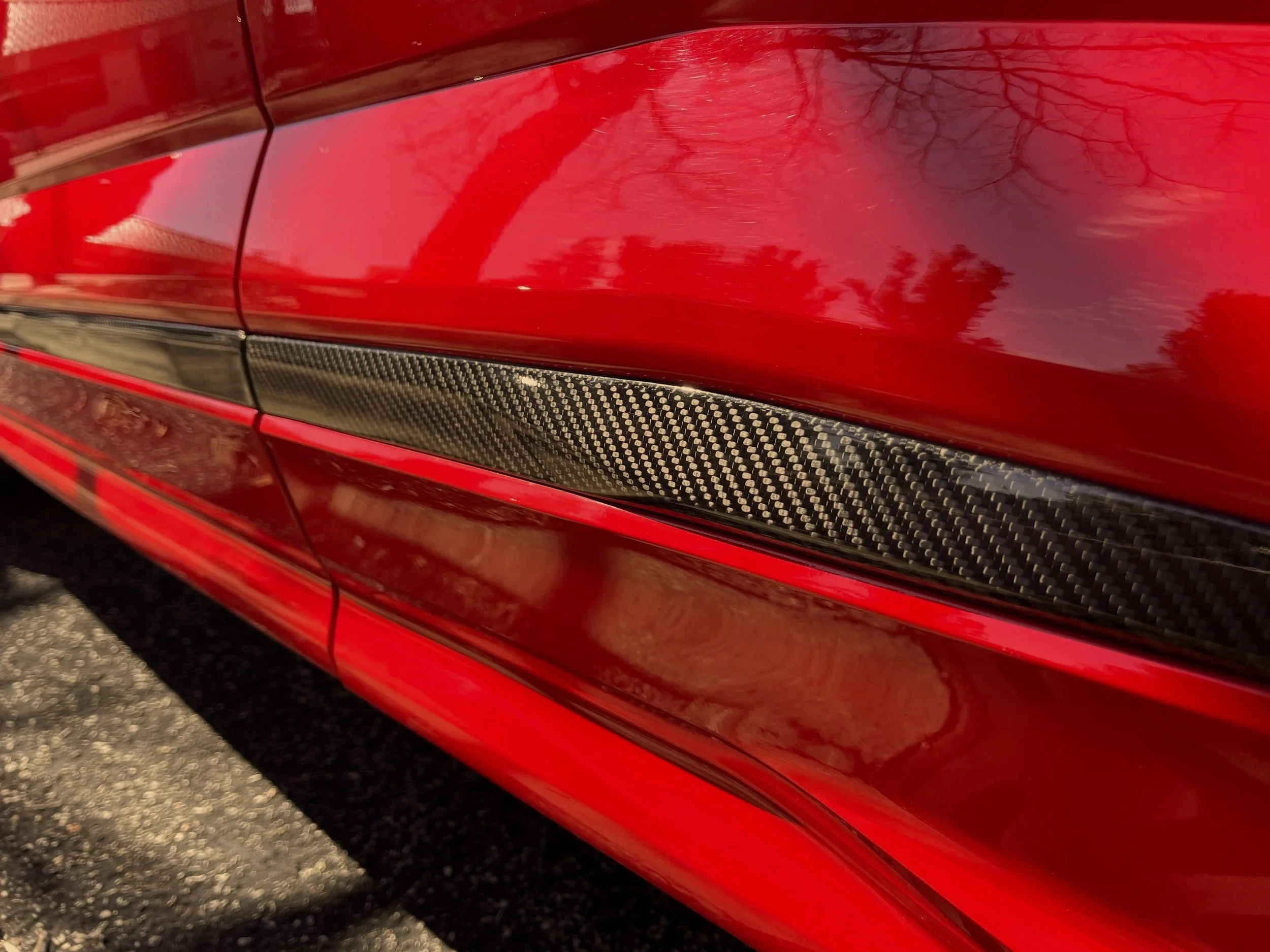 Close-up of a red car's side with a carbon fiber strip and visible scratches on the paint.