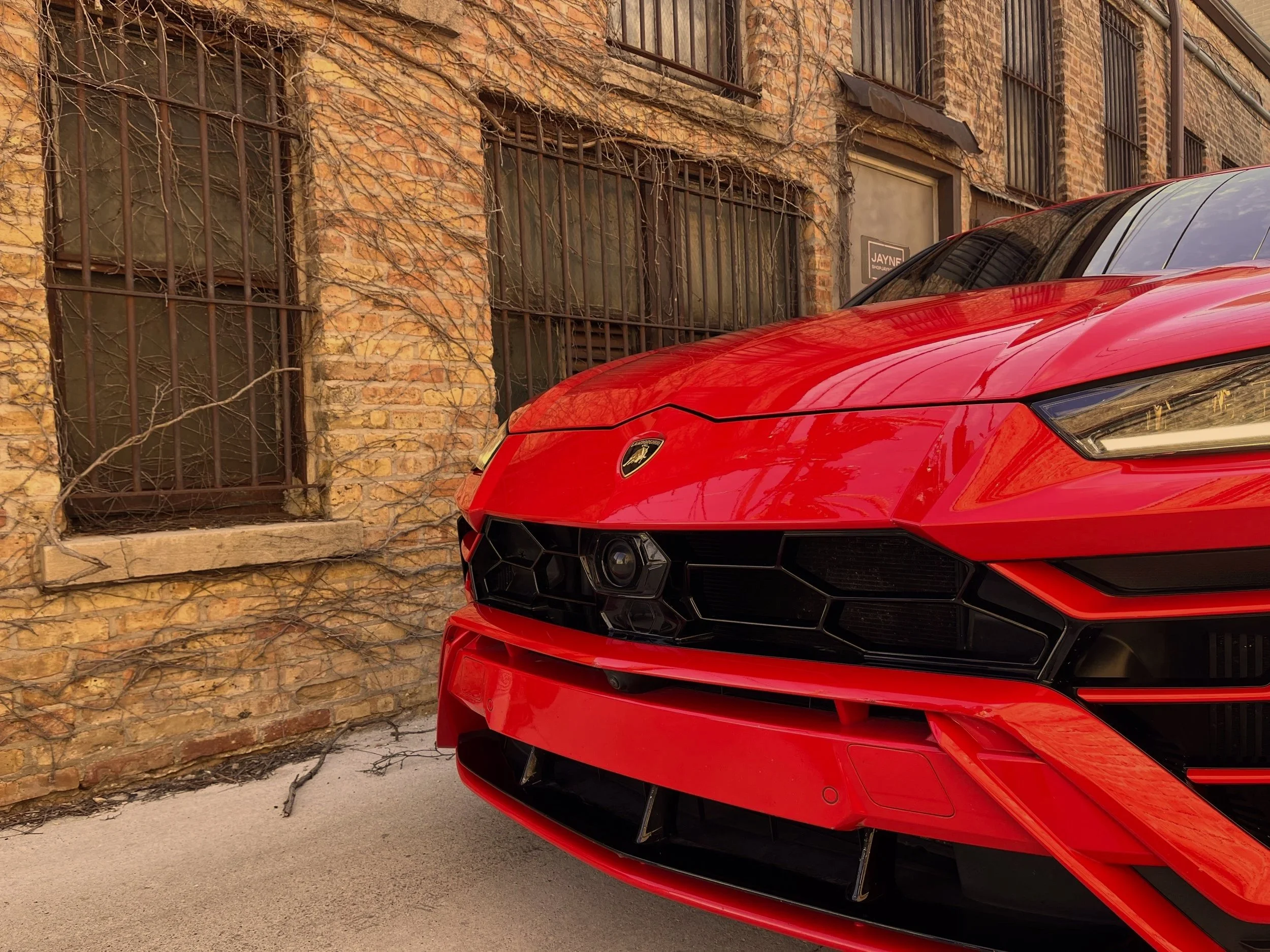 Close-up of a red Lamborghini car parked in front of a brick building with barred windows and dried vines crawling on the walls.