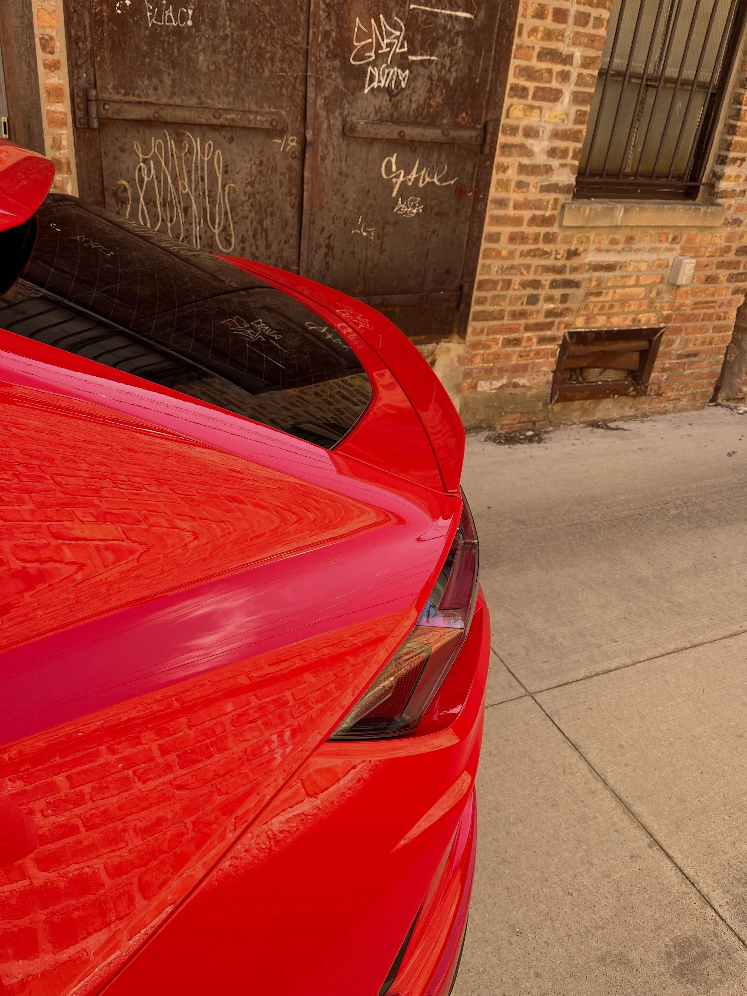 Close-up view of the rear part of a bright red car, showing the back window and part of the trunk, parked on a city street. In the background, an old rusty metal door with graffiti and a brick wall with a window are visible.
