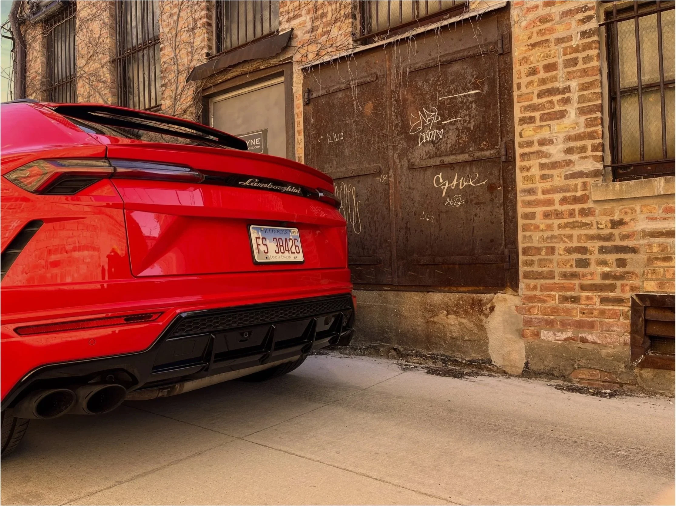 Red Lamborghini car parked beside a brick building with barred windows and a rusty metal door with graffiti. The license plate indicates Illinois registration.