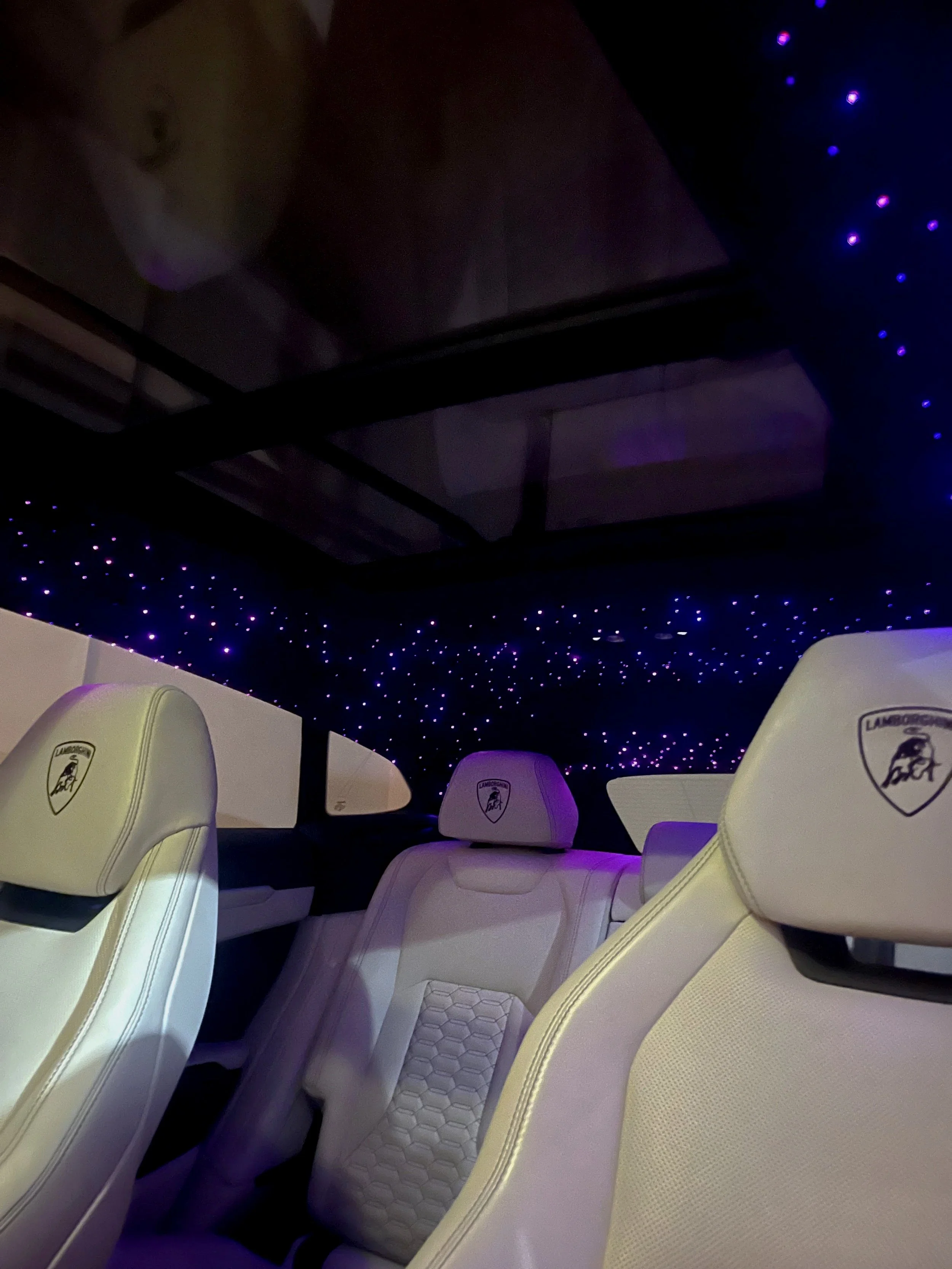Luxury white interior car seats with Lamborghini logo, black ceiling with purple LED star lights, and a panoramic glass roof.