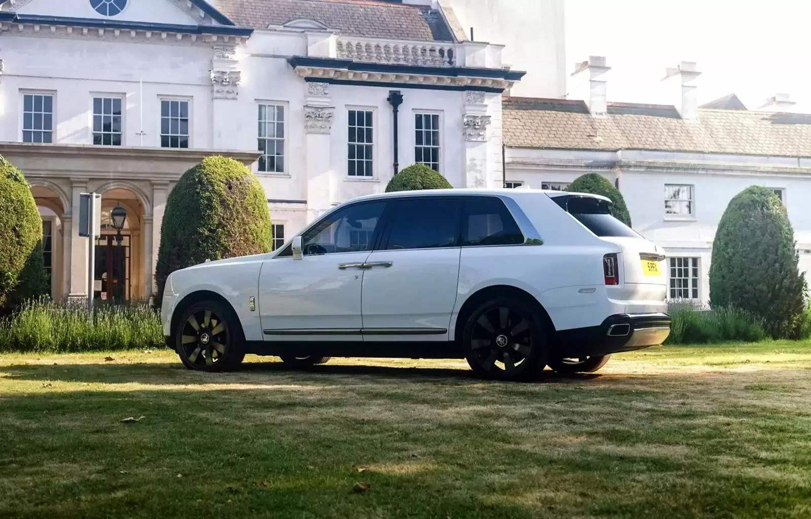 A white luxury SUV parked on a lawn in front of a large white house with tall windows and decorative architectural details.