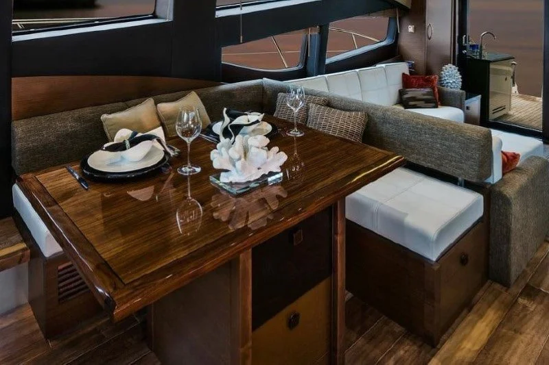 A wooden dining table set for two with a floral centerpiece and wine glasses, in a cozy dining area with sofas, cushions, and a window view.