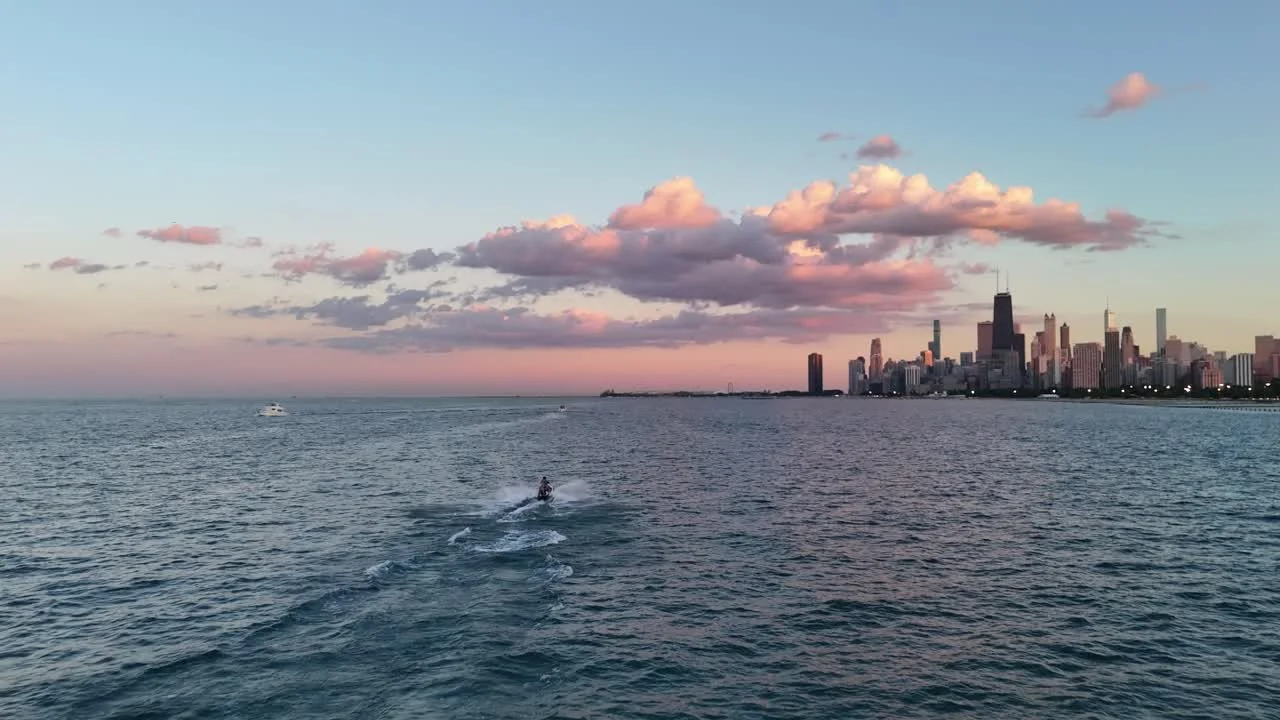 View of a city skyline across a body of water with a boat and a person riding a jet ski in the foreground during sunset or sunrise.