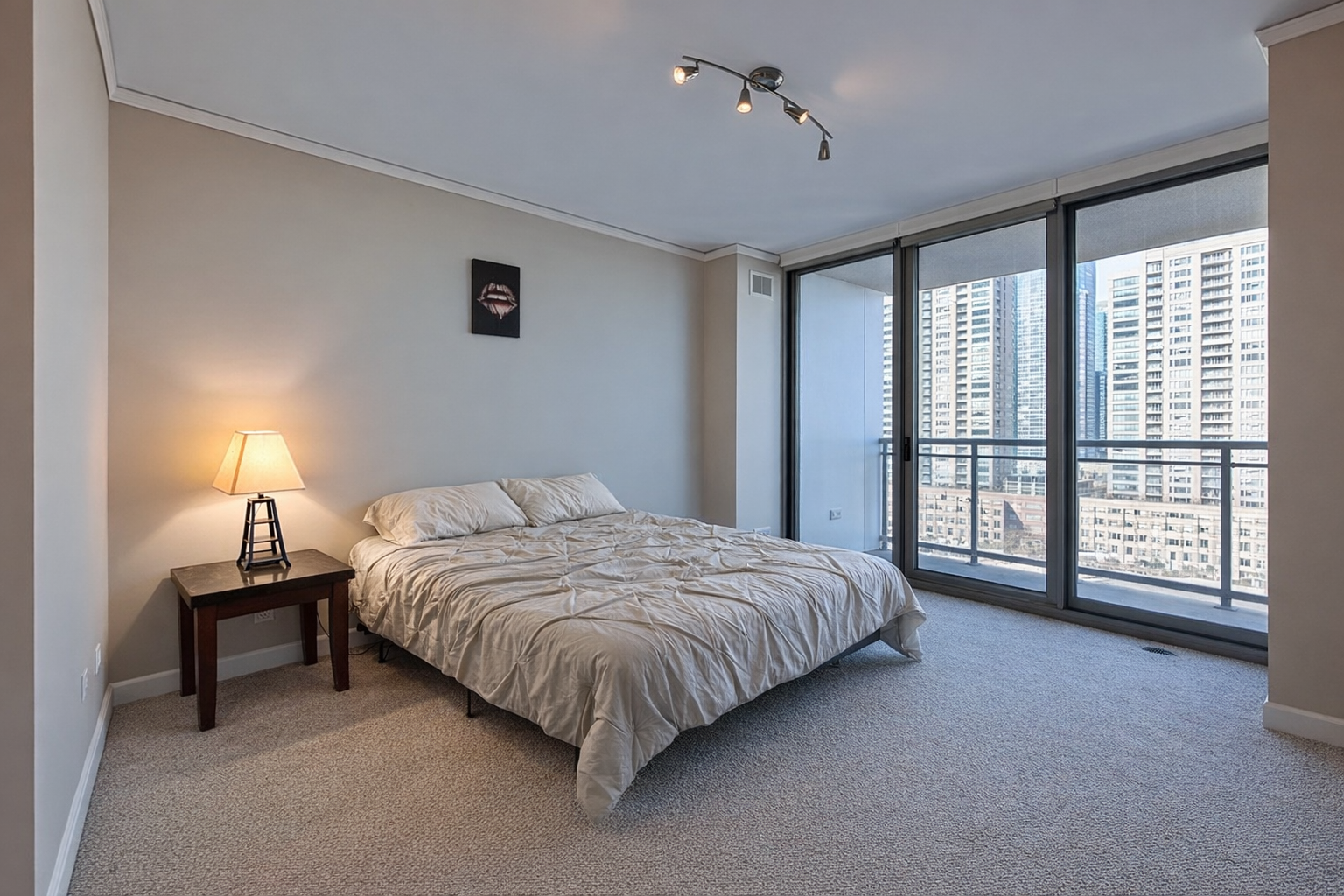 Bedroom with bed, nightstand with lamp, large windows showing city skyline.