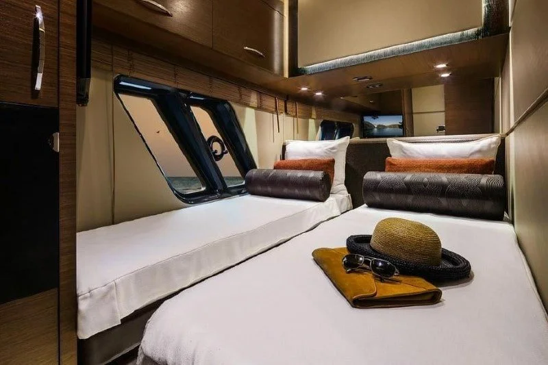 Two beds placed side by side in a small, cozy room with windows, pillows, and a tray holding a hat, sunglasses, and a notepad.