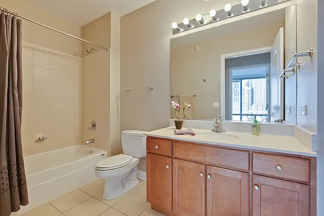 Bathroom with beige tile flooring, a bathtub with a shower, a white toilet, a wooden vanity with a sink, a large mirror, and a window with city view.