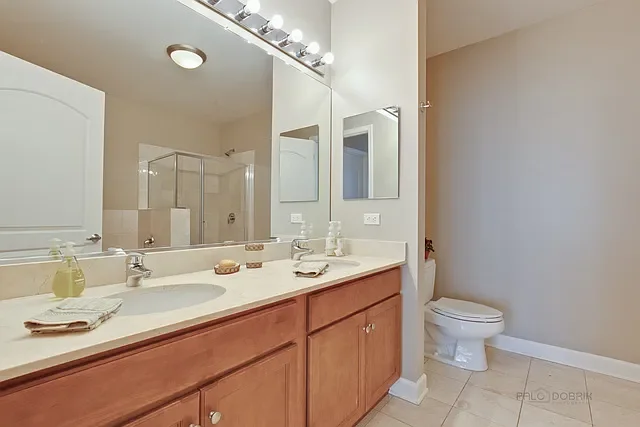 Bathroom with double vanity sink, large mirror, and a shower stall in the background. There is a toilet on the right side and a beige wall.