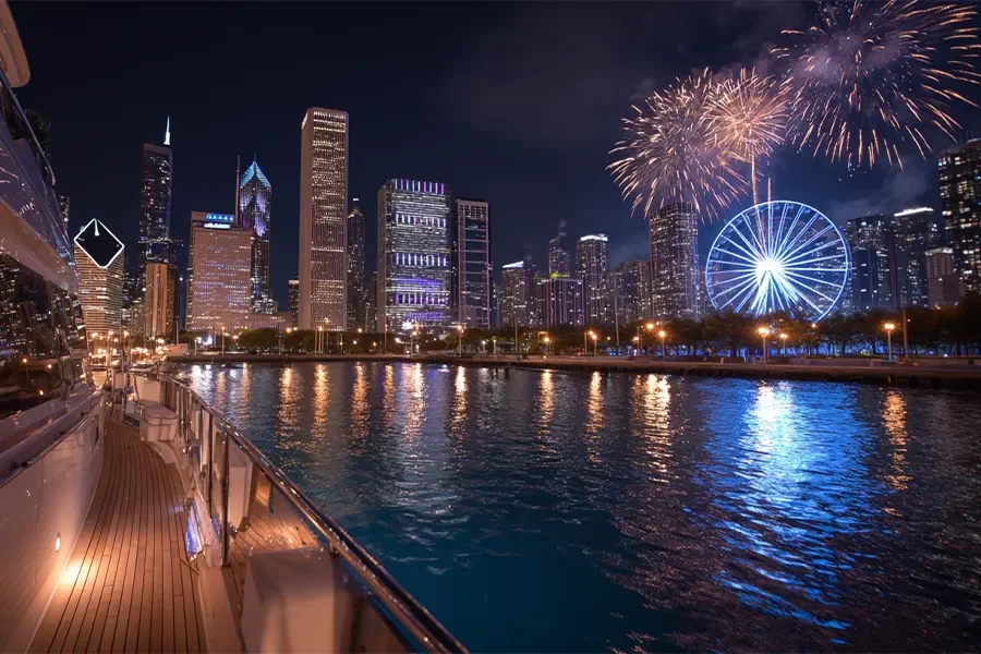 Nighttime city skyline with fireworks over a river, viewed from a boat docked along the water.