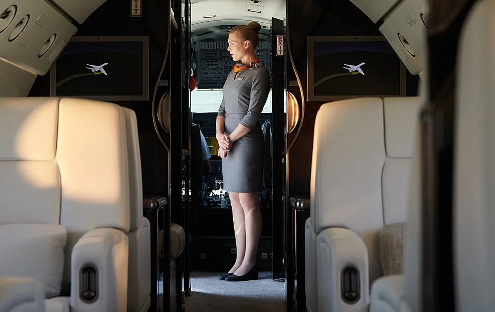Flight attendant standing inside an airplane cabin near the cockpit door, with beige seats and overhead compartments visible.