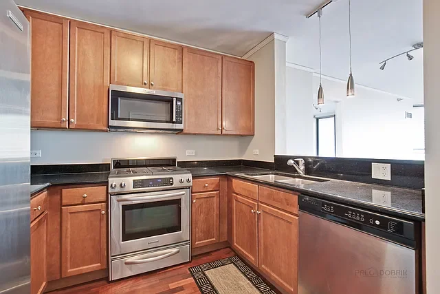 Kitchen with wooden cabinets, stainless steel microwave, oven, and dishwasher, black countertops, and a small area rug on hardwood floor.