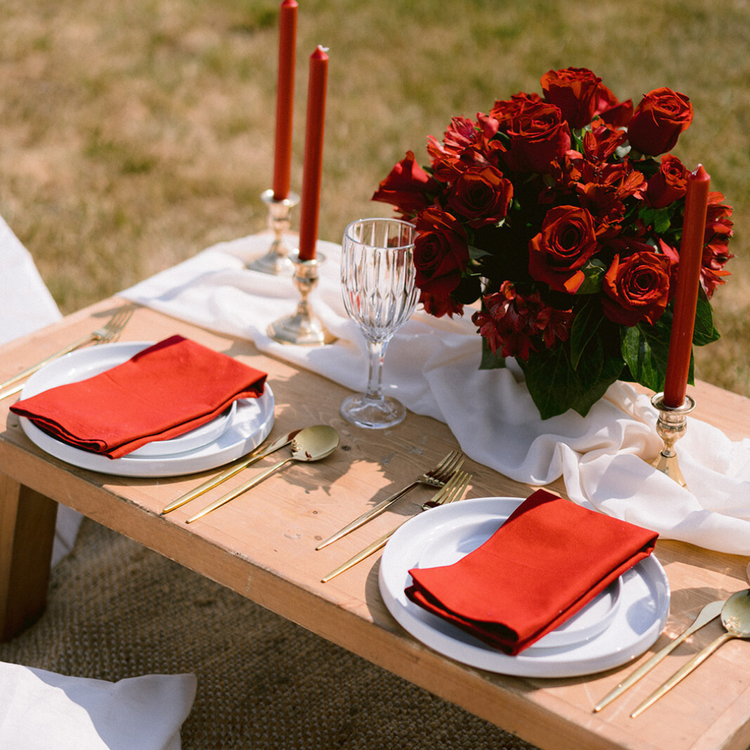 Table setting with white plates, red napkins, gold utensils, a floral centerpiece of red roses, two red candles, and an empty wine glass, outdoors on a grassy area.