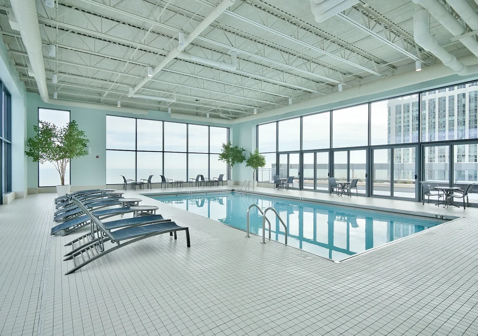 Indoor swimming pool area with large window walls, several lounge chairs lined up along the pool, tables and chairs around the perimeter, and potted trees, in a modern high-rise building.
