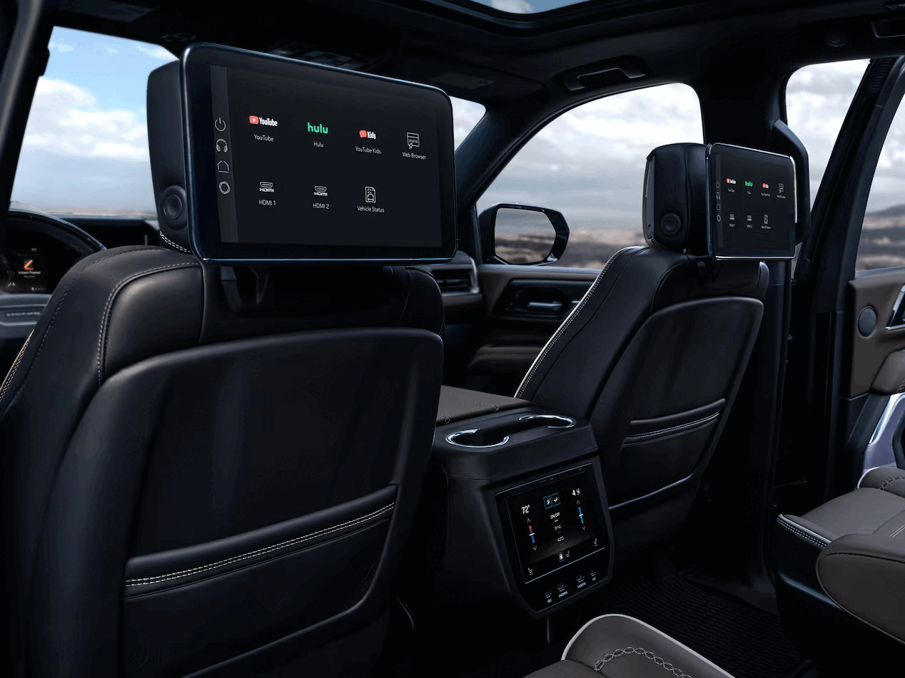Inside a modern vehicle showing two rear seat entertainment screens displaying streaming apps, air conditioning controls, and the landscape outside the front windshield with a partly cloudy sky.