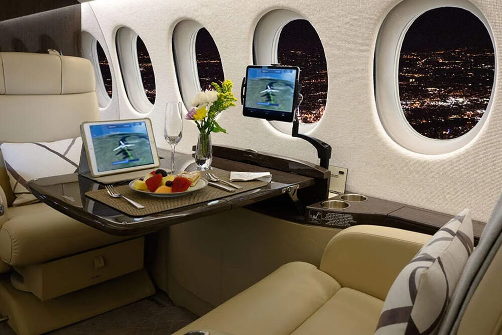 Luxury airplane interior with a dining setup, including a tablet, a glass of champagne, a bowl of fruit, and a flower arrangement, overlooking night city lights outside the windows.