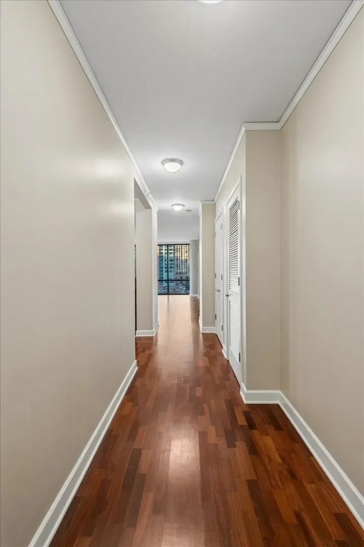Empty hallway with light-colored walls, white molding, hardwood floors, and ceiling lights leading to a balcony with city view.