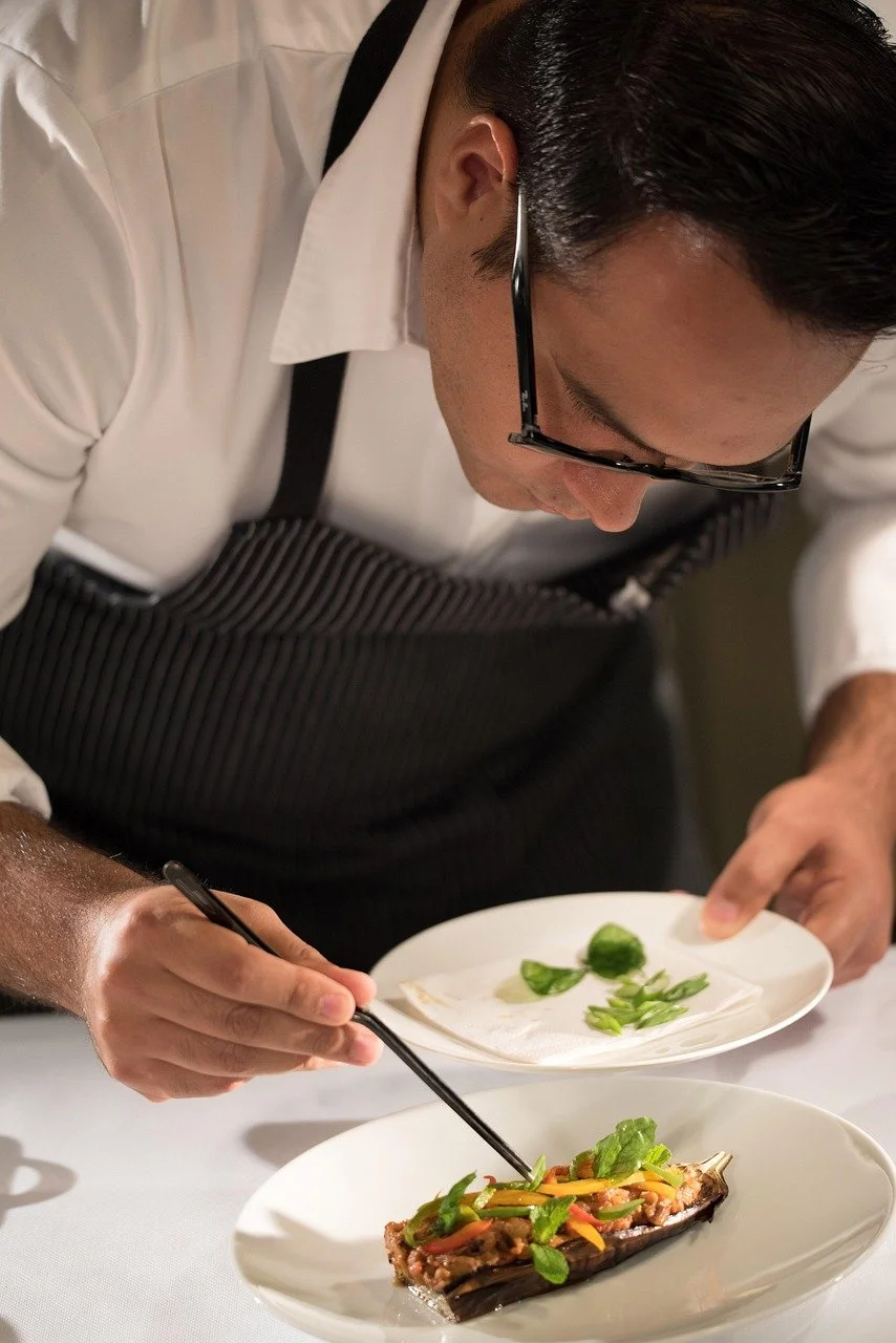 A chef garnishing a cooked fish with herbs and vegetables on a white plate.