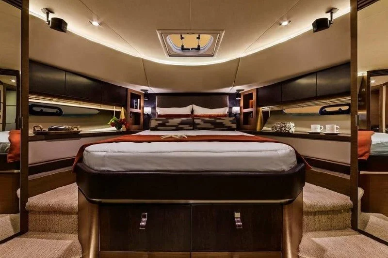 Luxurious bedroom with a large bed, pillows, and decorative pillows, illuminated by built-in lighting, with a ceiling window and side shelves holding decor and beverages.