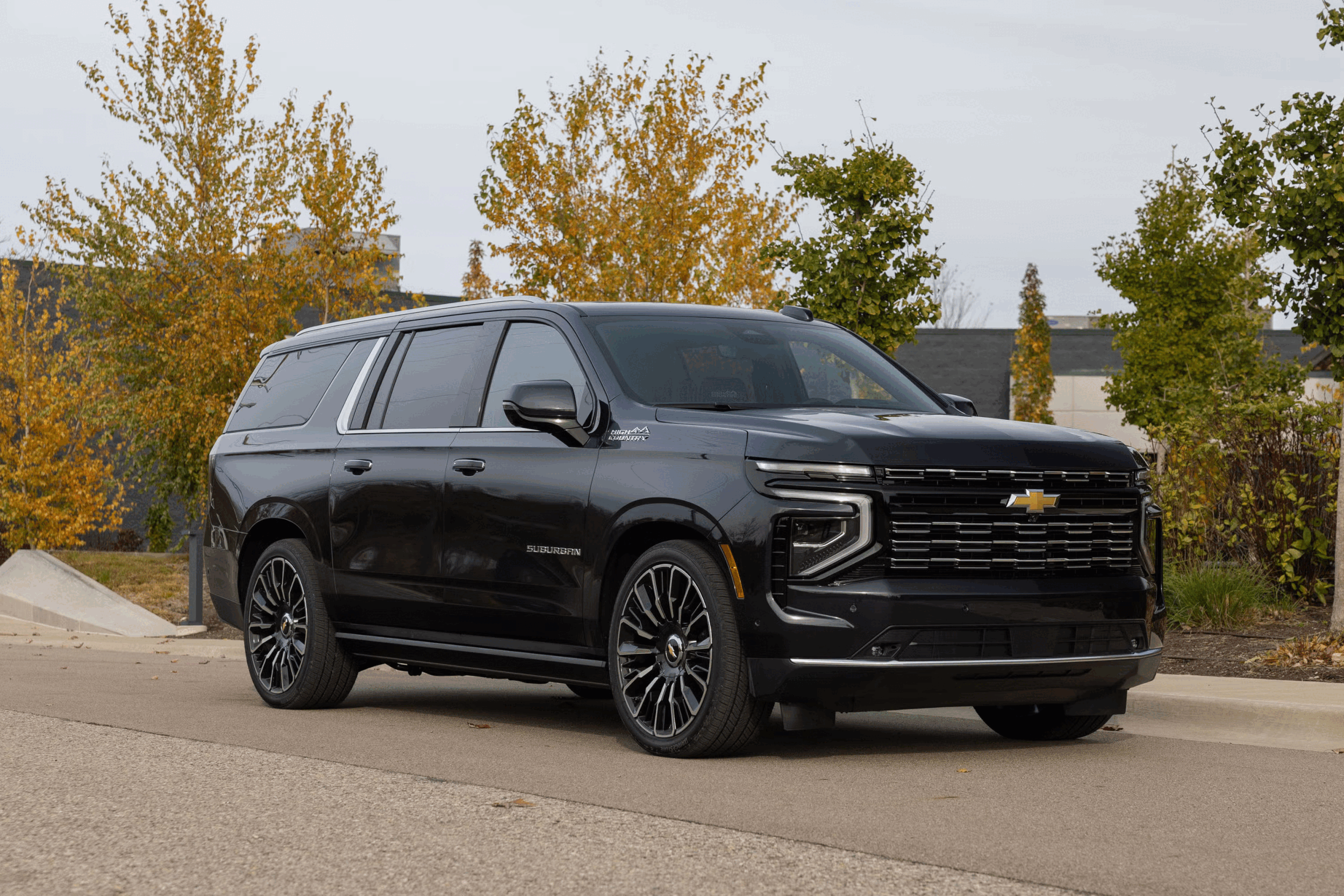 A black Chevrolet Suburban parked in a parking lot with autumn-colored trees and buildings in the background.