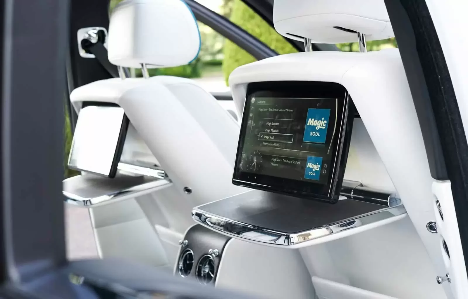 Inside the front seat area of a modern vehicle with a white interior, featuring a digital infotainment screen displaying a radio station called Magic Soul, and multiple screens and controls on the dashboard.