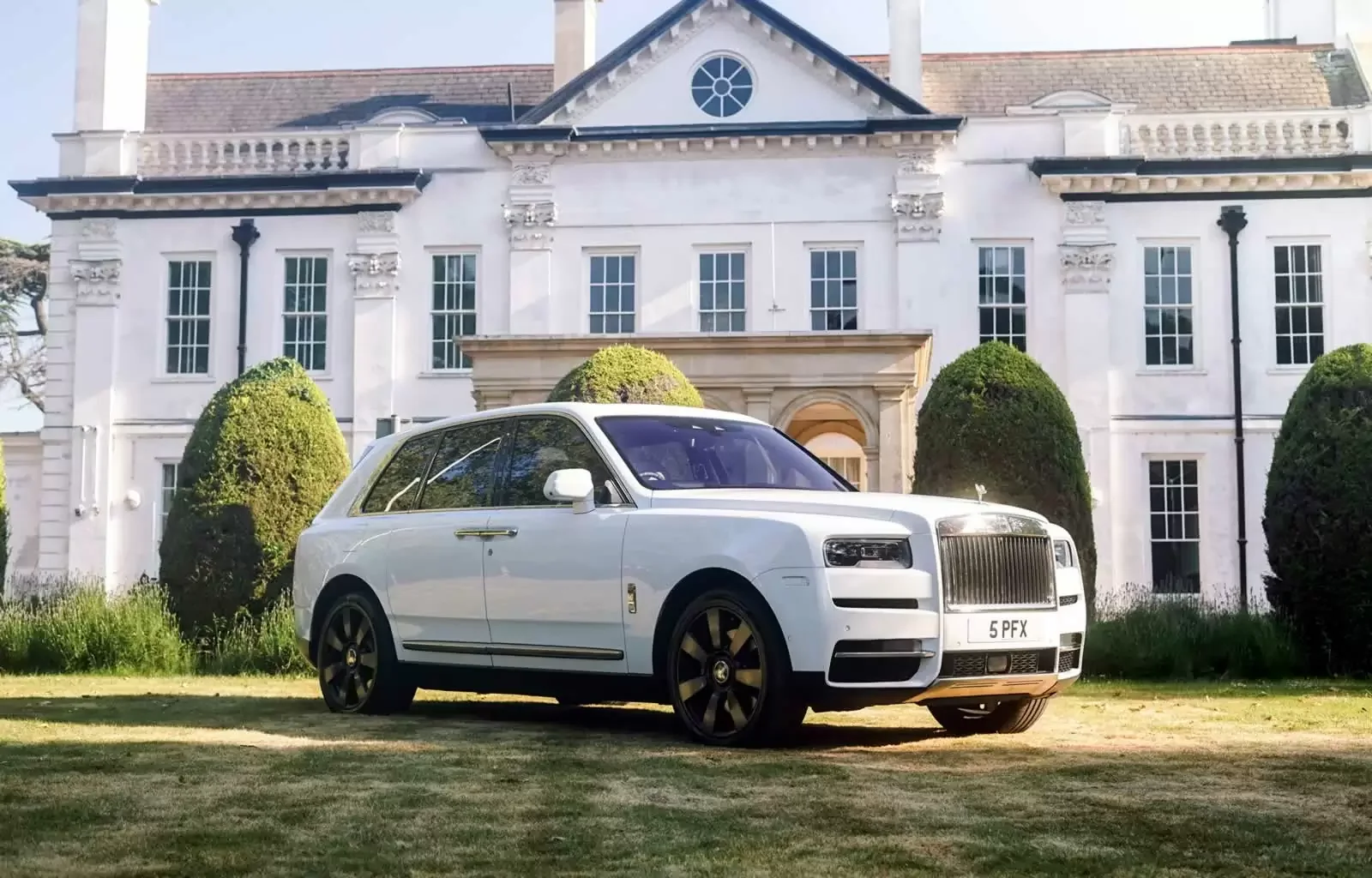 A white luxury SUV parked on a lawn in front of a large white mansion with multiple windows and neatly trimmed shrubs.