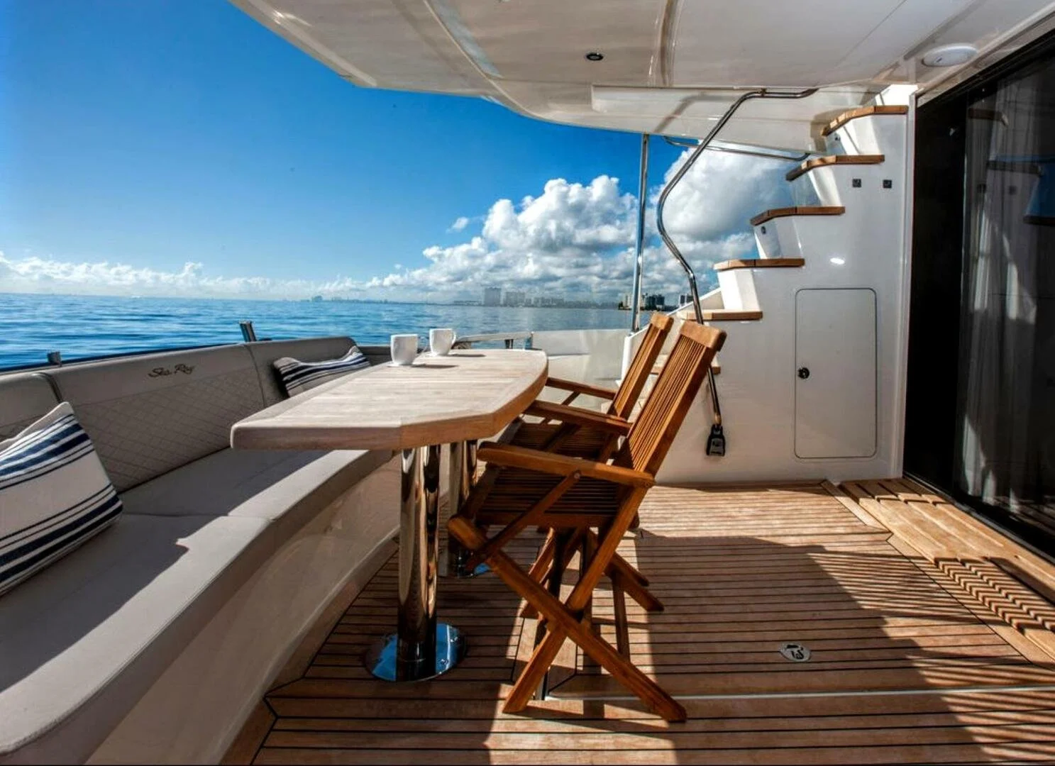 View of a yacht deck with a wooden table, chairs, and outdoor cushions, overlooking the ocean with blue sky and clouds