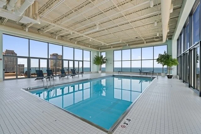Indoor swimming pool area with large windows, lounge chairs, and potted plants.