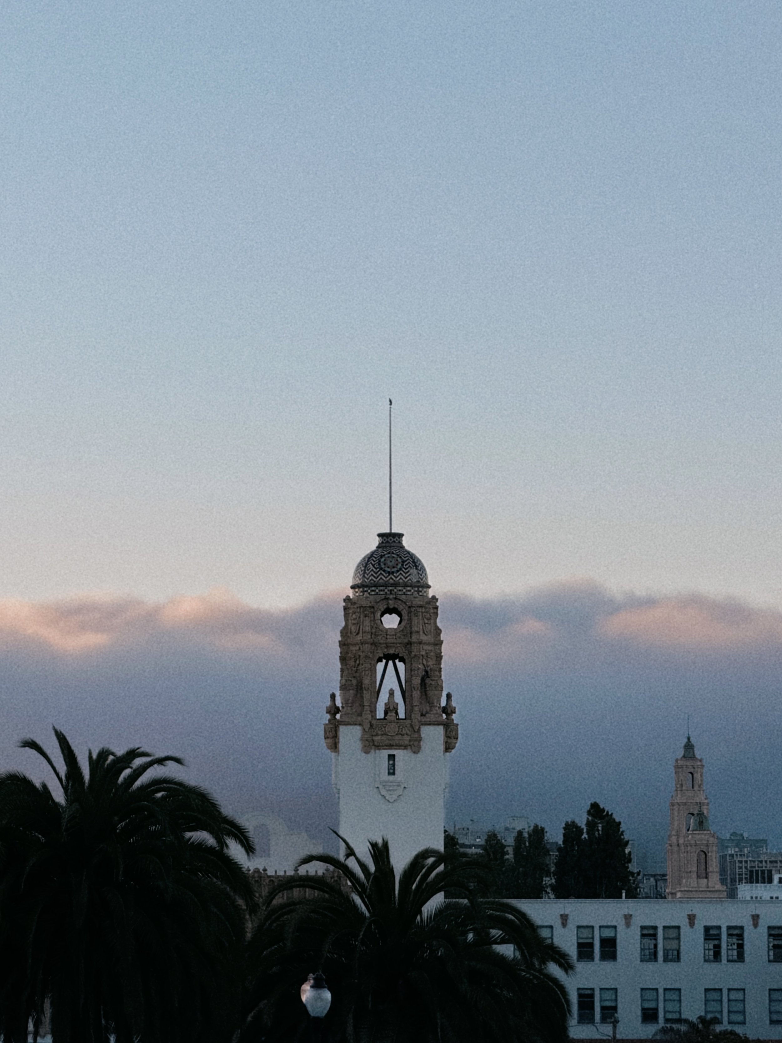 A clock tower with ornate architectural details, topped with a small dome and a flag, set against a sky at dusk with a few clouds and a cityscape in the background.
