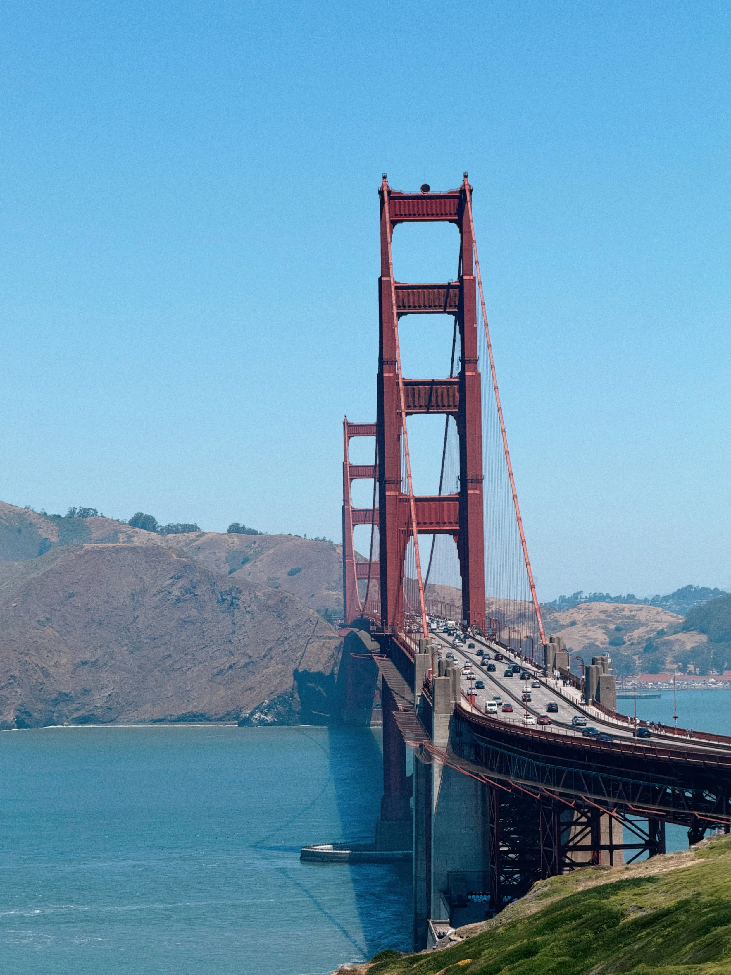 The Golden Gate Bridge extending over water with cars traveling across it, with hills in the background under a clear blue sky.