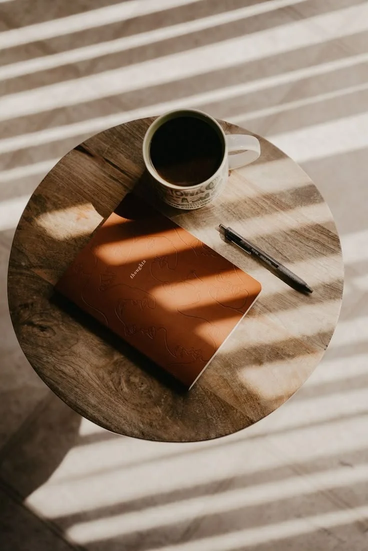 Taza de café sobre una mesa de madera, acompañada de un cuaderno marrón y un bolígrafo, con sombras de persianas en el fondo.