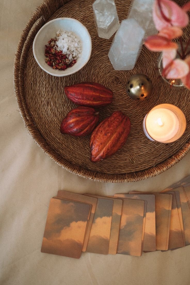 Vela encendida, tres frutas que parecen cacao fermentado, cristales de sal, pétalos de rosa, esfera dorada, cartas de tarot con ilustraciones de nubes y cielo, y flores en un florero en una mesa de mimbre.