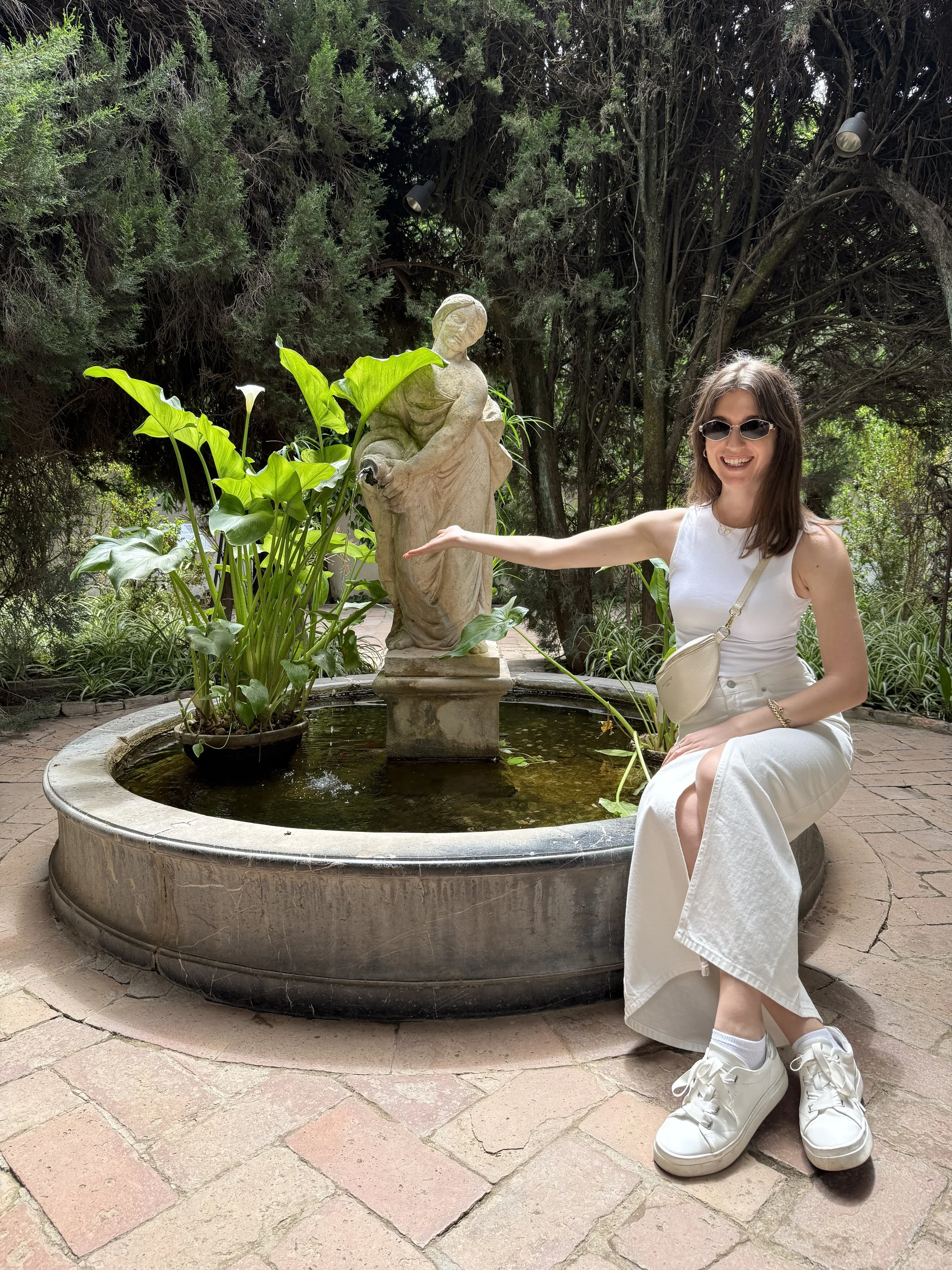A young woman with shoulder-length brown hair wearing sunglasses, a white sleeveless top, white skirt, white sneakers, and carrying a small beige purse kneeling beside a stone fountain. The fountain features a sculpture of a woman pouring water, surrounded by green foliage and tall trees in the background.