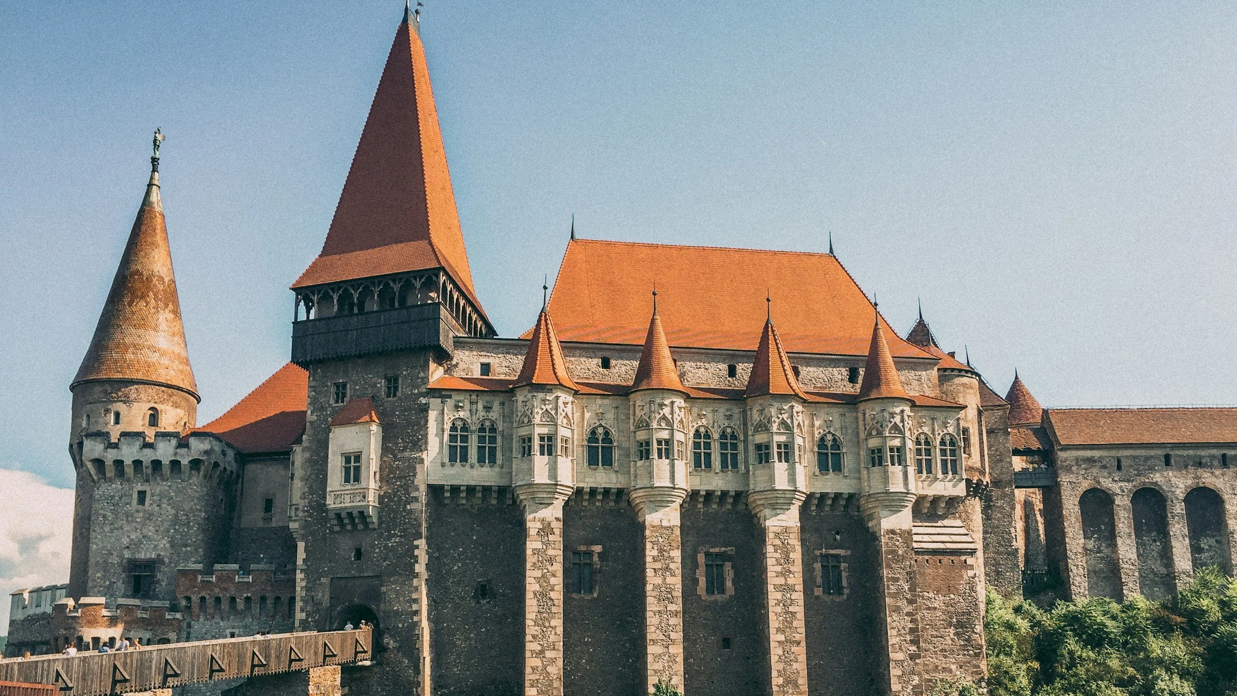View of a historic castle with stone walls, multiple towers with red-tiled roofs, and a blue sky in the background.