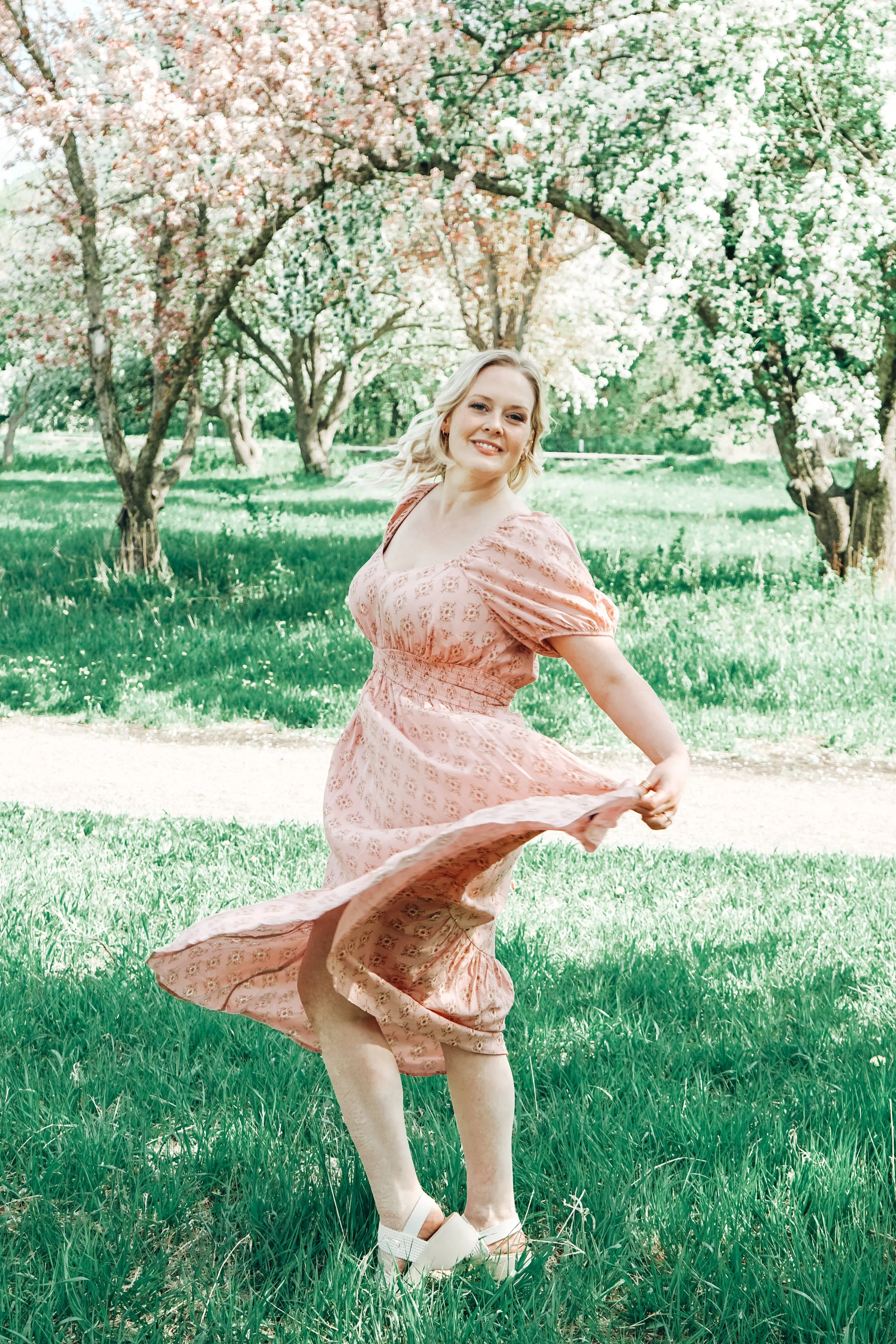 A woman in a pink floral dress twirls in a grassy park with blooming white and pink flowering trees in the background, smiling.