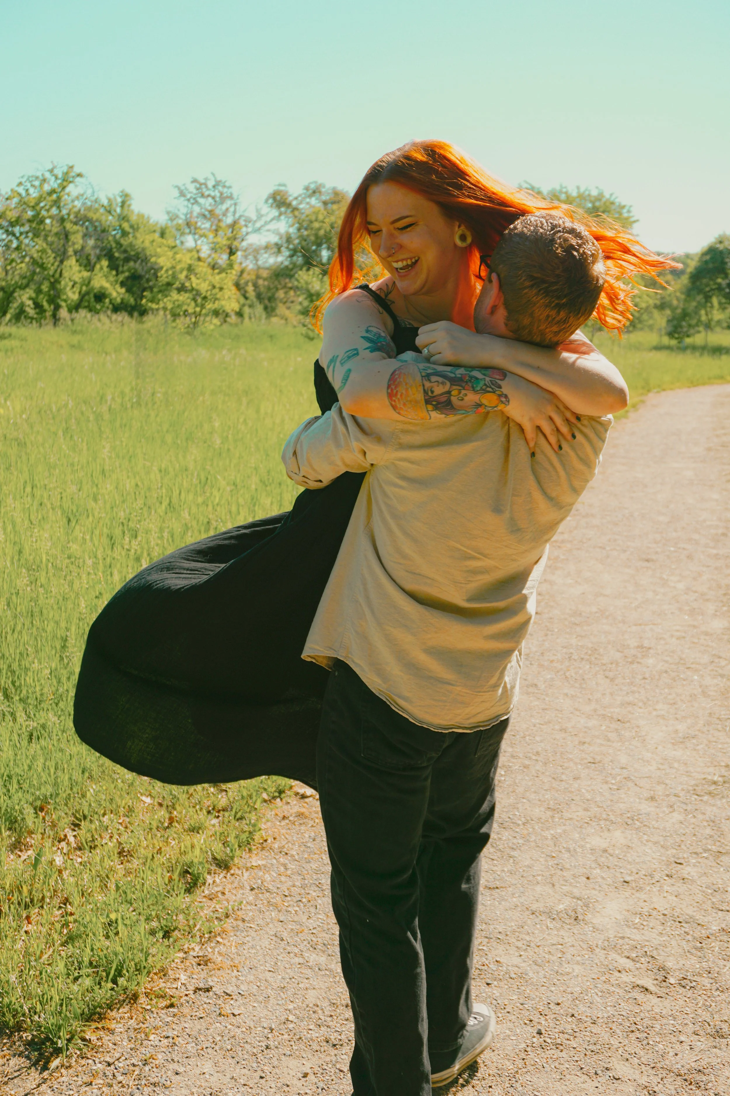 A couple outdoors on a sunny day, with the man joyfully carrying the woman, both smiling and embracing each other on a dirt path with green grass and trees in the background.