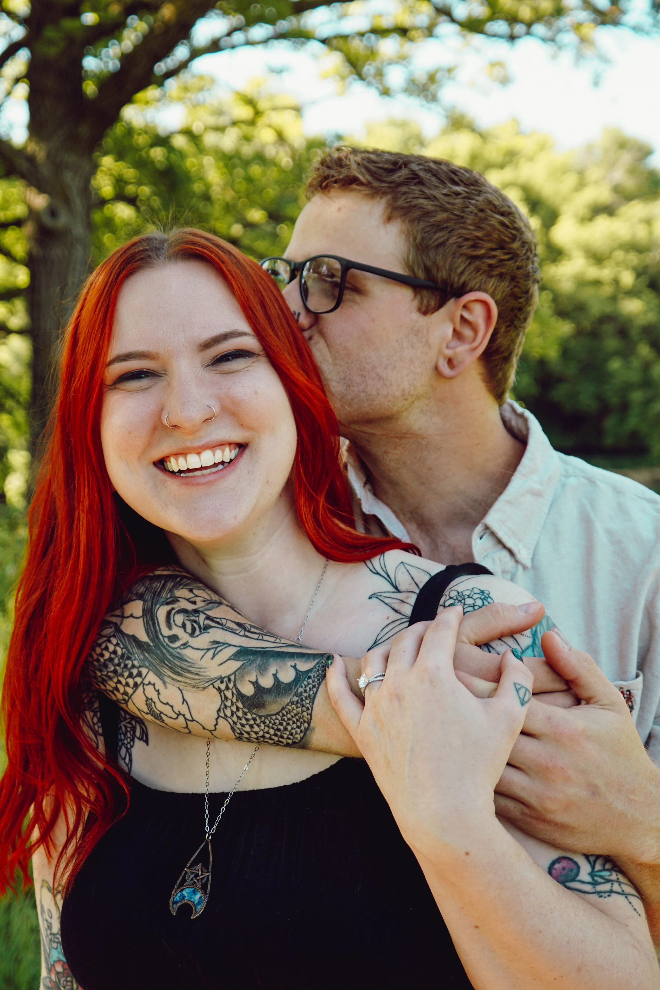 A smiling woman with long red hair, tattoos on her arms and chest, wearing a black top and jewelry, being kissed on the cheek by a man with glasses and short brown hair, in an outdoor setting with trees and sunlight.